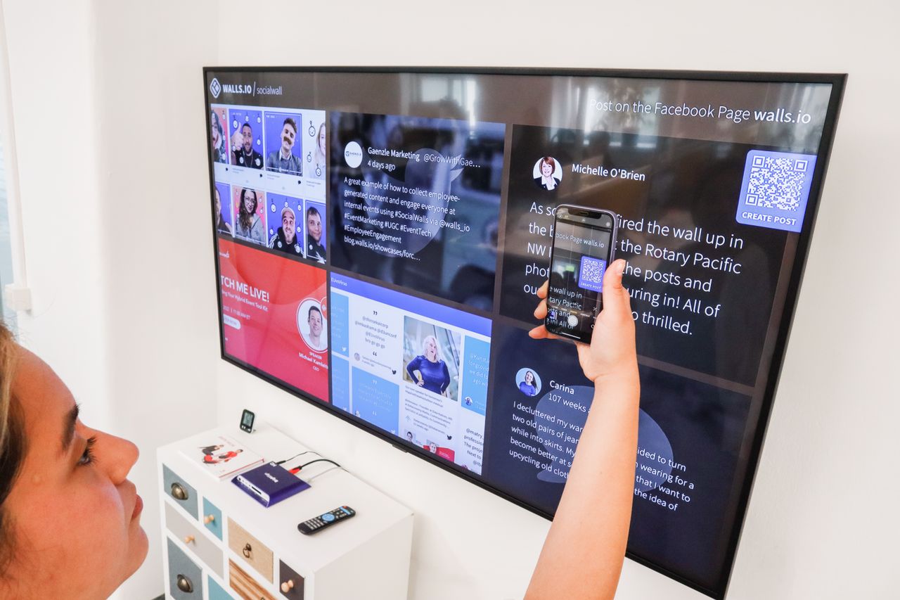 A woman is using a smartphone to scan a QR code shown on a digital wall display indoors.